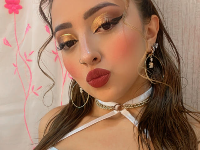 KylieCharm avec des lèvres rouges pulpeuses, un fard à paupières doré, des sourcils épais, de longs cheveux bruns ondulés, un collier blanc, de grandes boucles d'oreilles et un anneau de nez, regardant vers le bas..