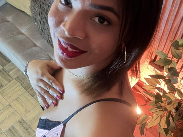AliceBooty with dark brown hair, red lipstick, and black lace bra, smiling at camera, hand on chest, pink top, gold necklace, hoop earrings, red nail polish, wooden floor.