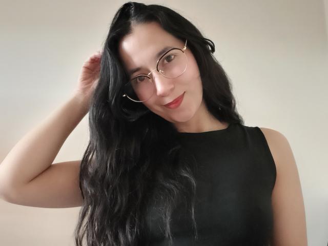 AnneStone with long black hair, wearing a black sleeveless top and round glasses, touching her hair with a subtle smile, showing her cute face and red lipstick.