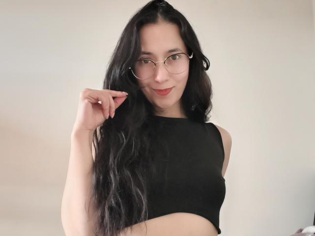 AnneStone with long black hair, glasses, and a black sleeveless top, pointing with her right hand, showing her bare midriff and a subtle smile.