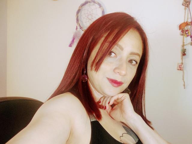 CatalinaMonyque with red hair, pale skin, pink lipstick, black tank top, and red nails, resting her chin on her hand, smiling subtly. Dreamcatcher and beads in the background.