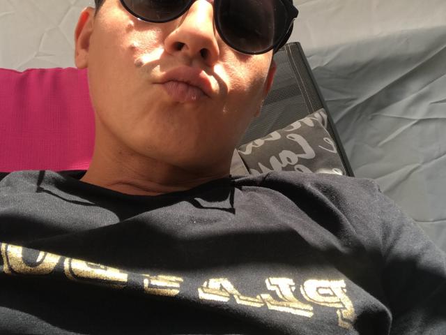 Tiz is lying down with dark sunglasses on, puffy lips in a kissy face, and a black shirt with gold letters. Sunlight highlights Tiz's light brown skin.