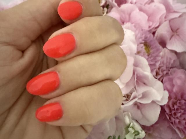 MargoRoiss's hand with bright red, glossy, almond-shaped nails, showing off shiny, well-maintained nails against a pink floral background.