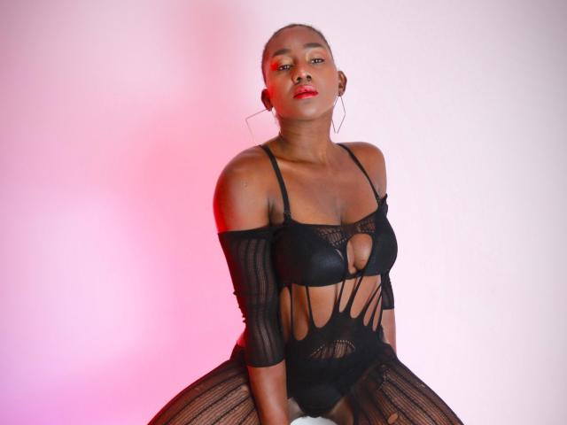 NahomyHawk with dark skin, shaved head, red lipstick, wearing black strappy lingerie that exposes her boobs and pussy, sitting against a pink and white gradient background.
