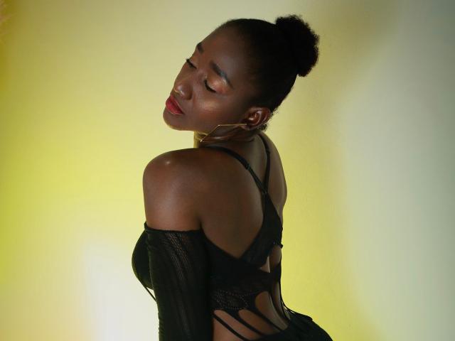 NahomyHawk with dark skin, red lipstick, and black lace lingerie, her back to the camera, head tilted, eyes closed, hair in a bun, wearing off-shoulder sleeves, looking sexy.