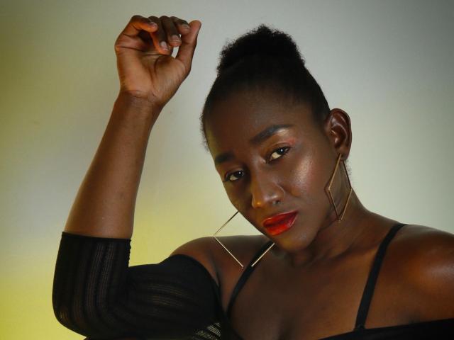 NahomyHawk, dark-skinned woman with red lipstick, raised arm, black off-shoulder top, gold hoop earrings, short curly hair, looking down with a slight smile, confident and sexy.