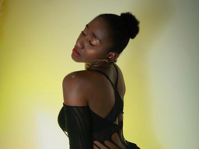 NahomyHawk with dark skin, eyes closed, red lipstick, black lace bra, and off-shoulder top, arms slightly raised, head tilted back, and hair in a bun, looking sexy.