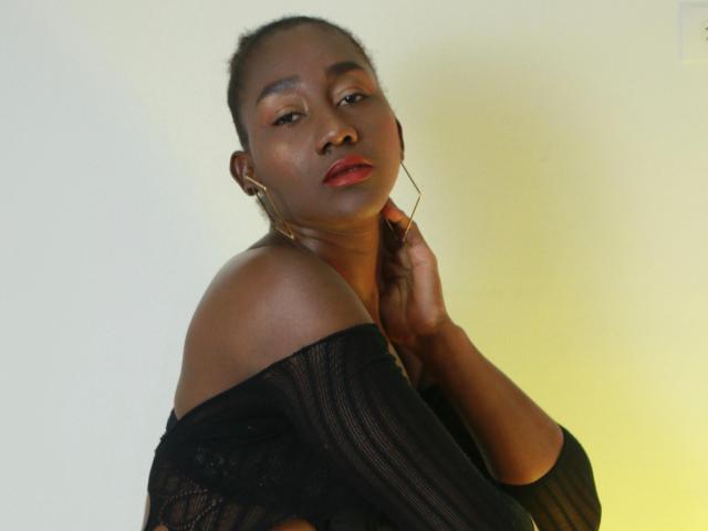NahomyHawk with dark skin, red lipstick, and black off-shoulder top, hand near neck, looking at camera, yellow light from side, earrings, serious expression.