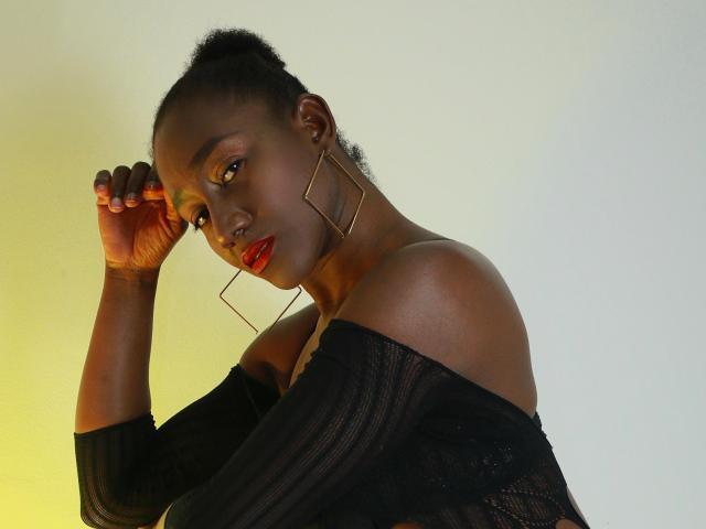 NahomyHawk, dark-skinned, with red lipstick, wearing a black off-shoulder top and gold geometric earrings, has a hand up, looking at the camera, in a yellow-lit photo.