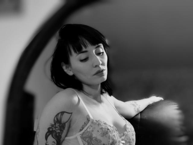 Black-and-white photo of SaraleePoison with short dark hair, eyes closed, wearing lace lingerie, arm resting on leather chair, showing a tattoo on her upper arm. Boobs visible.