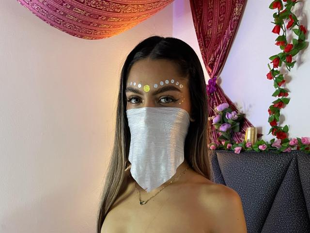 DalilaAliX with brown hair, wearing a silver mask, white face dots, and necklace, exposing bare shoulders, looking at camera. Boobs visible, no visible pussy. Decorated room.