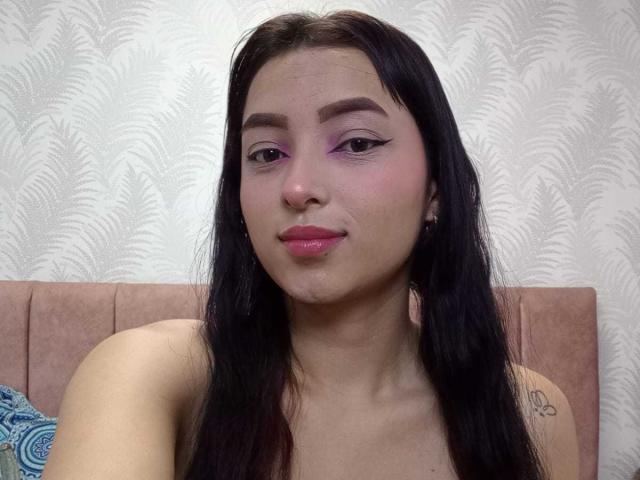 MaraLewis has long black hair, pink eyeliner, and pink lipstick, with a tattoo on her right shoulder, and is looking at the camera with a slight smile.