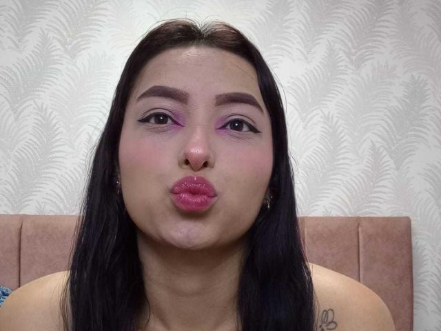 MaraLewis with pink eyeshadow and purple eyeliner, puffy pink lips, black hair, slightly chubby face, small tits, small tattoo on right shoulder, pink headboard, pouty expression.