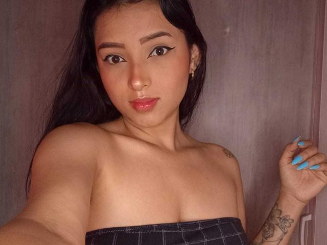 MaraLewis with dark hair, brown eyes, and blue nails, wearing a strapless black top, showing her tits, nose ring, tattoo on her right arm, and a subtle smile.