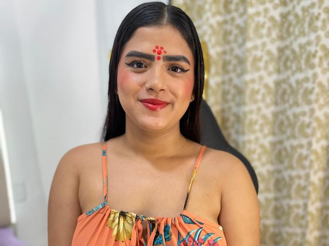NayaYalitza, with dark straight hair, red dot on forehead, wearing an orange floral dress, smiling with red lipstick, brown eyes, and black eyeliner. Her tits are small.