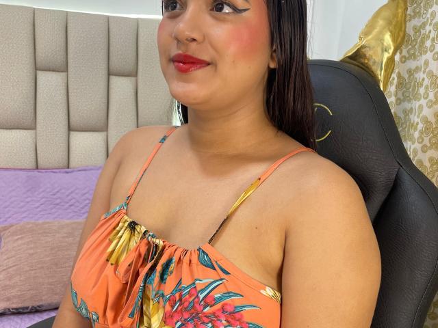 NayaYalitza with medium tits, brown skin, red lipstick, black eyeliner, and a smile, wearing an orange floral dress, sitting in a black chair.