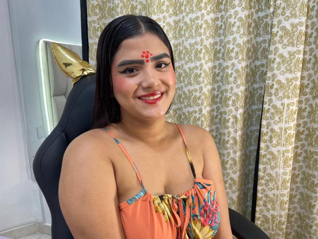 NayaYalitza with medium brown skin, black hair, red dot on forehead, smiling, wearing orange dress with colorful pattern, black chair, gold ear, and patterned curtain in background.