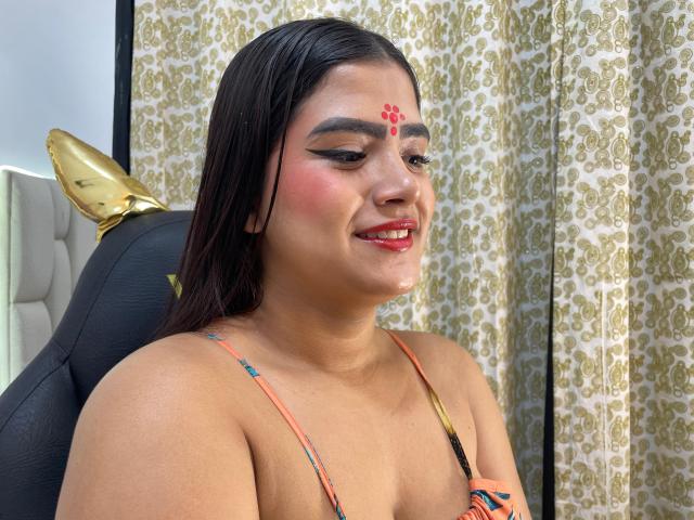 NayaYalitza has medium tits, brown skin, and a red bindi on her forehead. She's smiling with red lipstick, wearing a colorful halter top, and has dark wet hair.