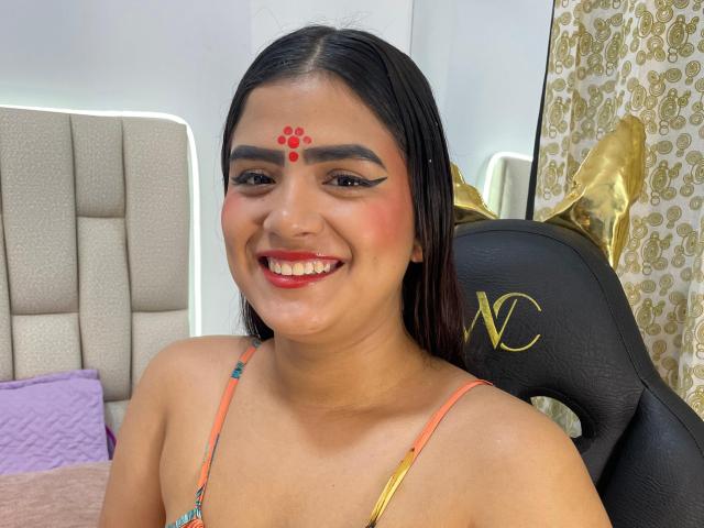 NayaYalitza with dark hair, red dot on forehead, black eyeliner, smiling, wearing colorful strap dress, black chair with gold "YC", pink pillow, gold decor in background.