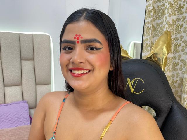 NayaYalitza has dark hair, red dot on forehead, black eyeliner, red lipstick, smiling, wearing colorful top, sitting in black chair with gold "NC" logo, tan skin, visible boobs.
