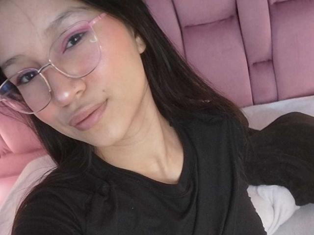 SophieDelaney with pink eyeshadow, clear glasses, black shirt, and long black hair, smiling slightly, lying on pink bed, soft light, gentle expression, subtle makeup, casual selfie.
