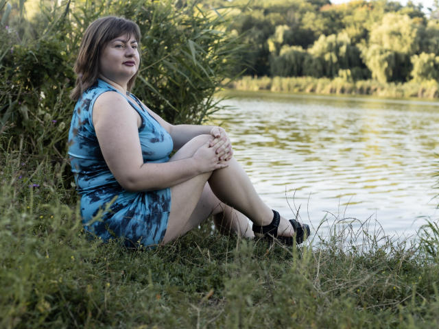 ZannaDreil with medium tits, blue tie-dye dress, sitting on grass by a lake, knees up, hands on knees, black heels, brown hair, smiling, green foliage in background.