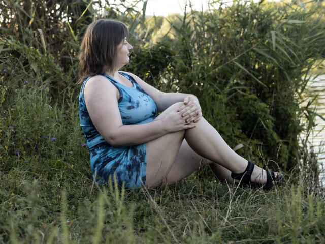 ZannaDreil, a chubby woman with brown hair, sits on grass wearing a blue tie-dye dress and black heels, legs bent, hands on knees, looking to the side.