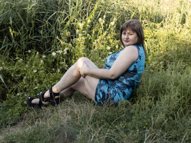 ZannaDreil sits in grass, legs bent, wearing a blue tie-dye dress, black heels, and showing her ass. She has brown hair, fair skin, and a slight smile.