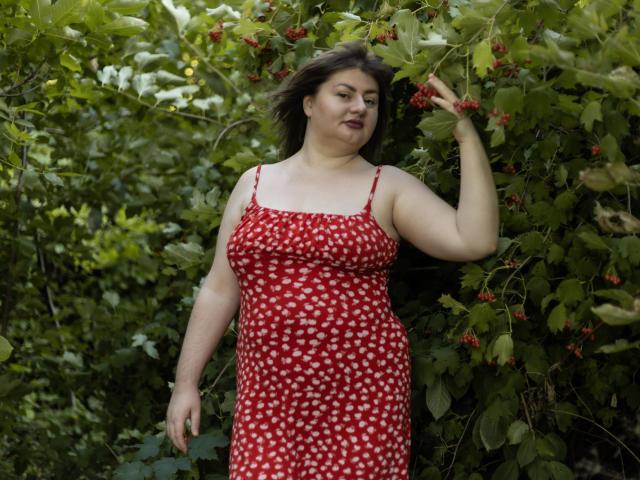ZannaDreil, plus-size with pale skin, brown hair, wearing a red dress with white hearts, standing in greenery, touching red berries, smiling slightly, right arm raised.