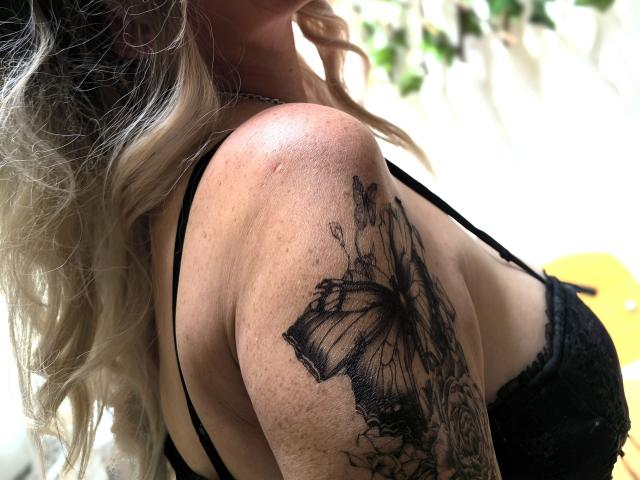 BigBoobsy69's upper arm tattoo of a large black butterfly with detailed wings and surrounding flowers, visible in a black lace bra, showing light wavy blonde hair.