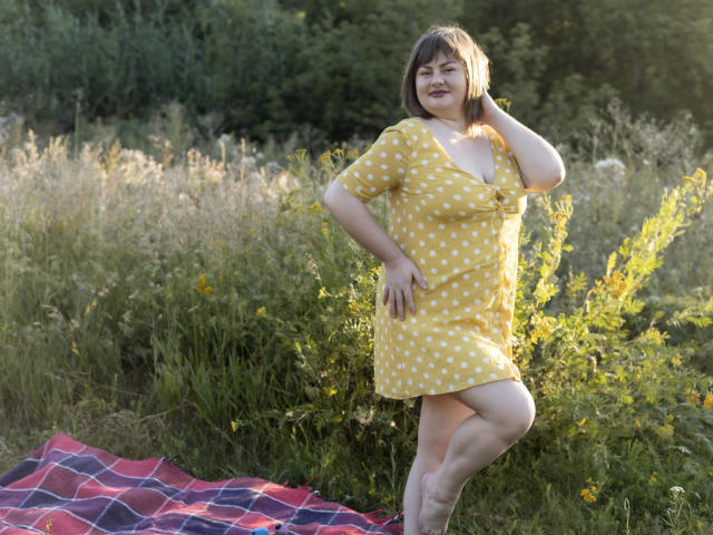 ZannaDreil stands in a grassy field, wearing a yellow polka dot dress, showing her tits, with one hand on her head and a smile, on a red plaid blanket.