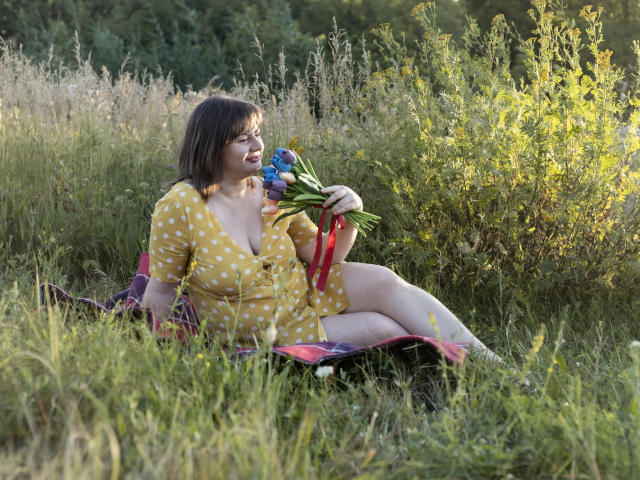 ZannaDreil with medium tits, yellow polka dot dress, sitting on grass holding blue flowers, smiling, brown bob haircut, legs crossed, purple blanket, daytime, greenery, sunlight.
