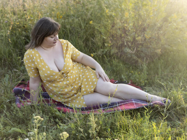 ZannaDreil with big boobs, pale skin, and brown hair, wearing a yellow polka dot dress, sitting on a red plaid blanket in a grassy field, looking down.