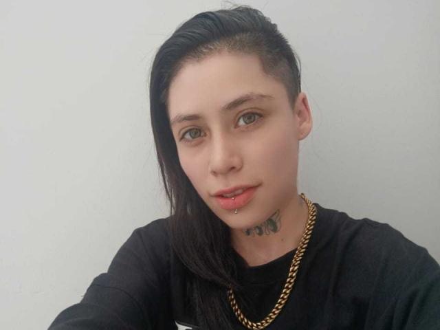 MorganWallen a une partie de cheveux rasée sur le côté, de longs cheveux noirs, une chaîne en or, un piercing aux lèvres et un tatouage de papillon sur le cou, et il porte un t-shirt noir..