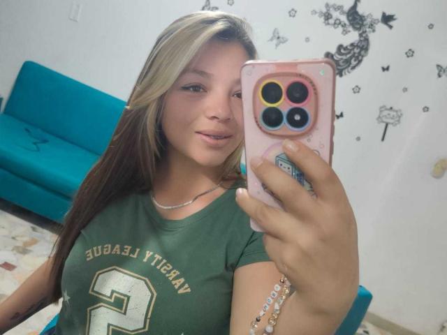 ScarlettAmelia with blonde and brown hair, green t-shirt, holding pink phone with colorful camera, smiling, showing ass, in selfie. Teasy, cute, and attractive.