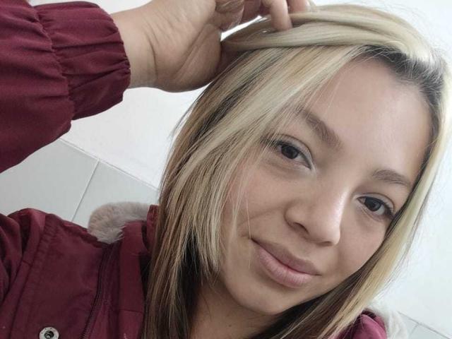 ScarlettAmelia with blonde and brown hair, smiling softly, wearing a red jacket, touching her head with one hand, pale skin, brown eyes, and subtle makeup.