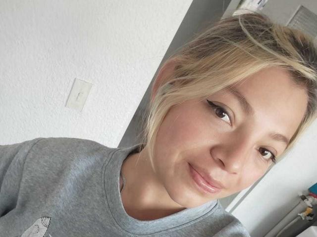 ScarlettAmelia has blonde hair in a messy bun, light skin, and is smiling softly at the camera, wearing a gray shirt, with brown eyes and subtle makeup.