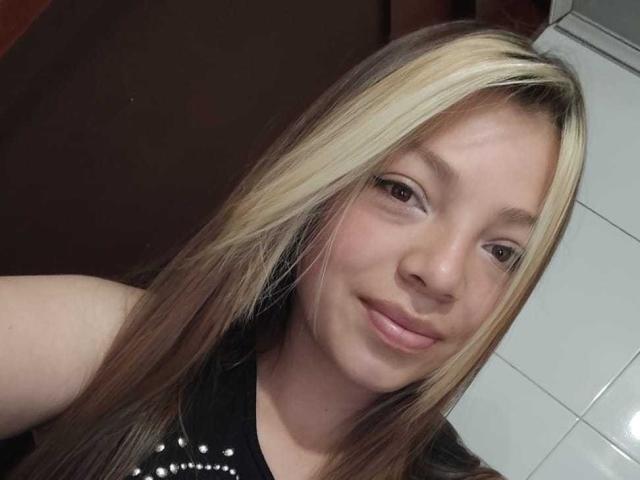 ScarlettAmelia has light brown skin, long blonde and brown hair, small nose, and a slight smile. She's wearing a black top with white studs, showing her ass.