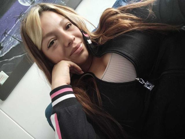 ScarlettAmelia, with long brown and blonde hair, leans on her hand, wearing a black zip-up jacket, showing her white bra-covered tits. She has a slight smile.
