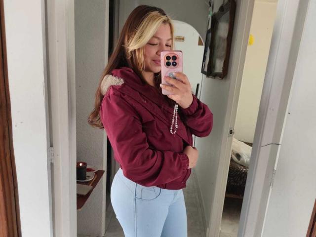 ScarlettAmelia with blonde and brown hair, wearing a maroon jacket and light blue jeans, holding a pink phone, showing her ass in a mirror selfie.