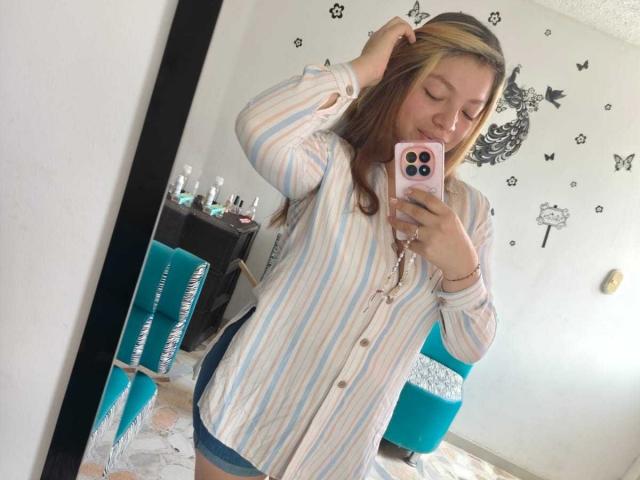 ScarlettAmelia with blonde highlights, wearing striped button-up shirt, blue shorts, holding pink phone, taking mirror selfie, hand in hair, cute, bedroom, visible bed and teal chair.