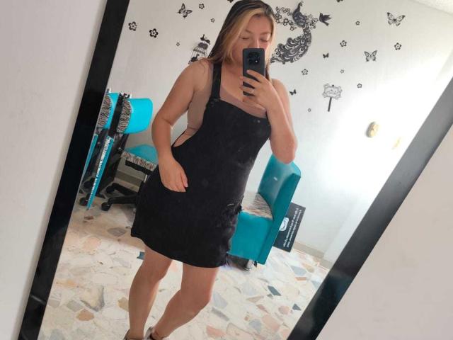 ScarlettAmelia in a black, sleeveless, lace dress, showing her ass, standing in front of a mirror, holding a phone, with blonde hair and blue eyes.