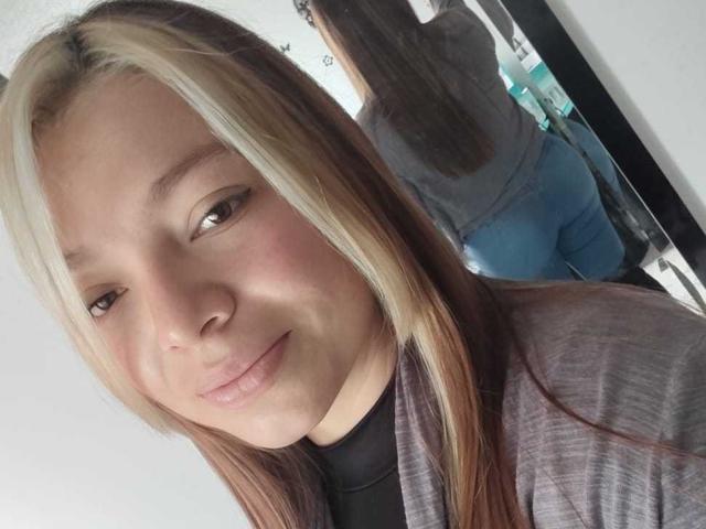 ScarlettAmelia has light brown and blonde hair, fair skin, and is smiling slightly. She's wearing a gray shirt, and her reflection shows her blue jeans and ass.