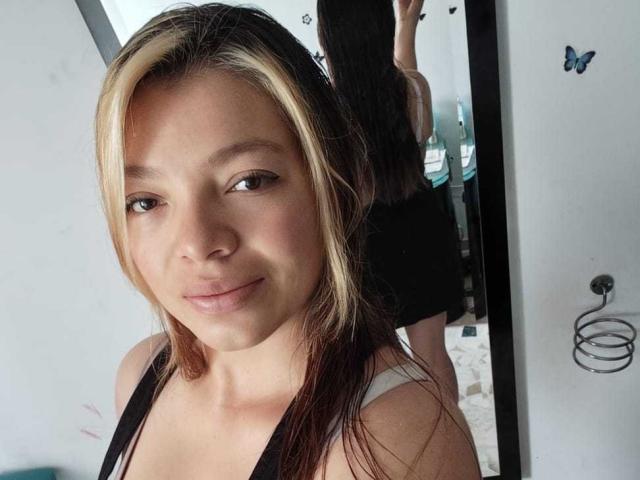 ScarlettAmelia has light brown skin, blonde and brown hair, brown eyes, small nose, and a slight smile. She's wearing a black and white top, reflecting in a mirror.