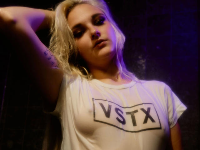 Scarlet-Young-hot with blonde hair, wearing a wet white T-shirt showing her tits, arm raised, purple and blue lighting, slightly annoyed expression, tattoo on upper arm.