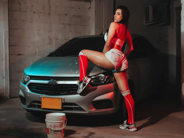 YelineMourine with big ass, wearing red knee socks and white shorts, leans on a silver car in a dim garage, her back to the camera, looking over her shoulder.