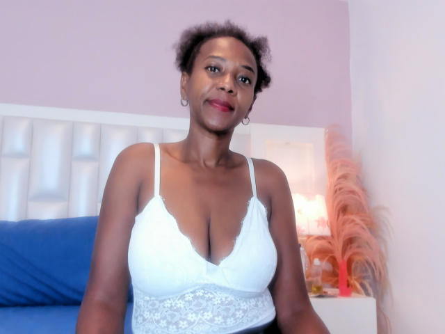 AfroHeat with dark skin, curly hair, and red lipstick, wearing a white lace bra that highlights her large, rounded tits, standing in a room with blue bedding.