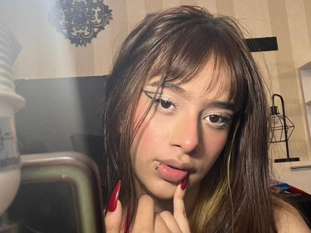 CamiMonet with brown eyes, dark brown hair, and red nails, biting her finger, showing a lip piercing, looking at a mirror. Her ass is not visible.