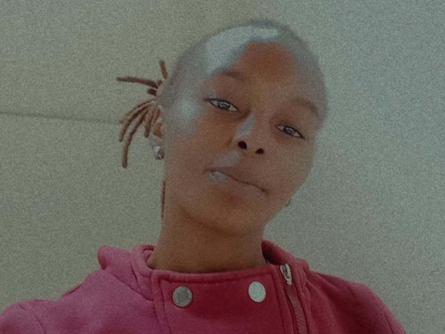 MollieClaudia with a bald head, brown skin, and small nose, wearing a pink hoodie with metal buttons, looking slightly to the side with a subtle smile.