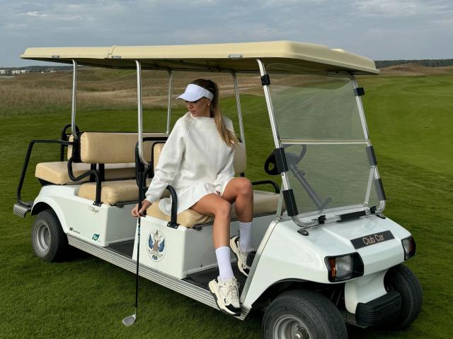 EngelEva-hot sits in a white golf cart, wearing a white visor, sweater, shorts, socks, and white shoes, holding a golf club, with green grass in the background.